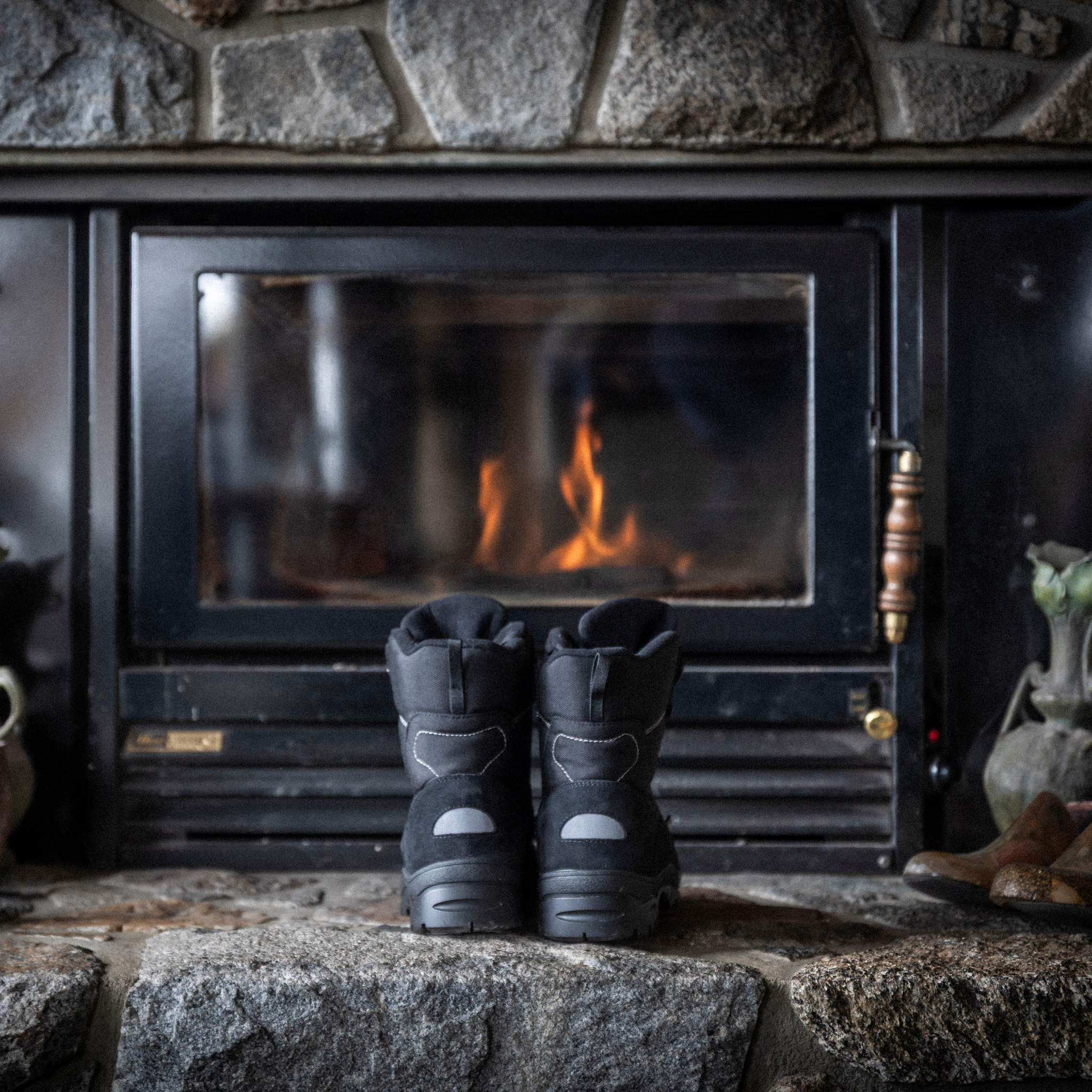 Pair of black Attiba Eiger 2 men's snow boots warming by a fireplace, rear three-quarter lifestyle view