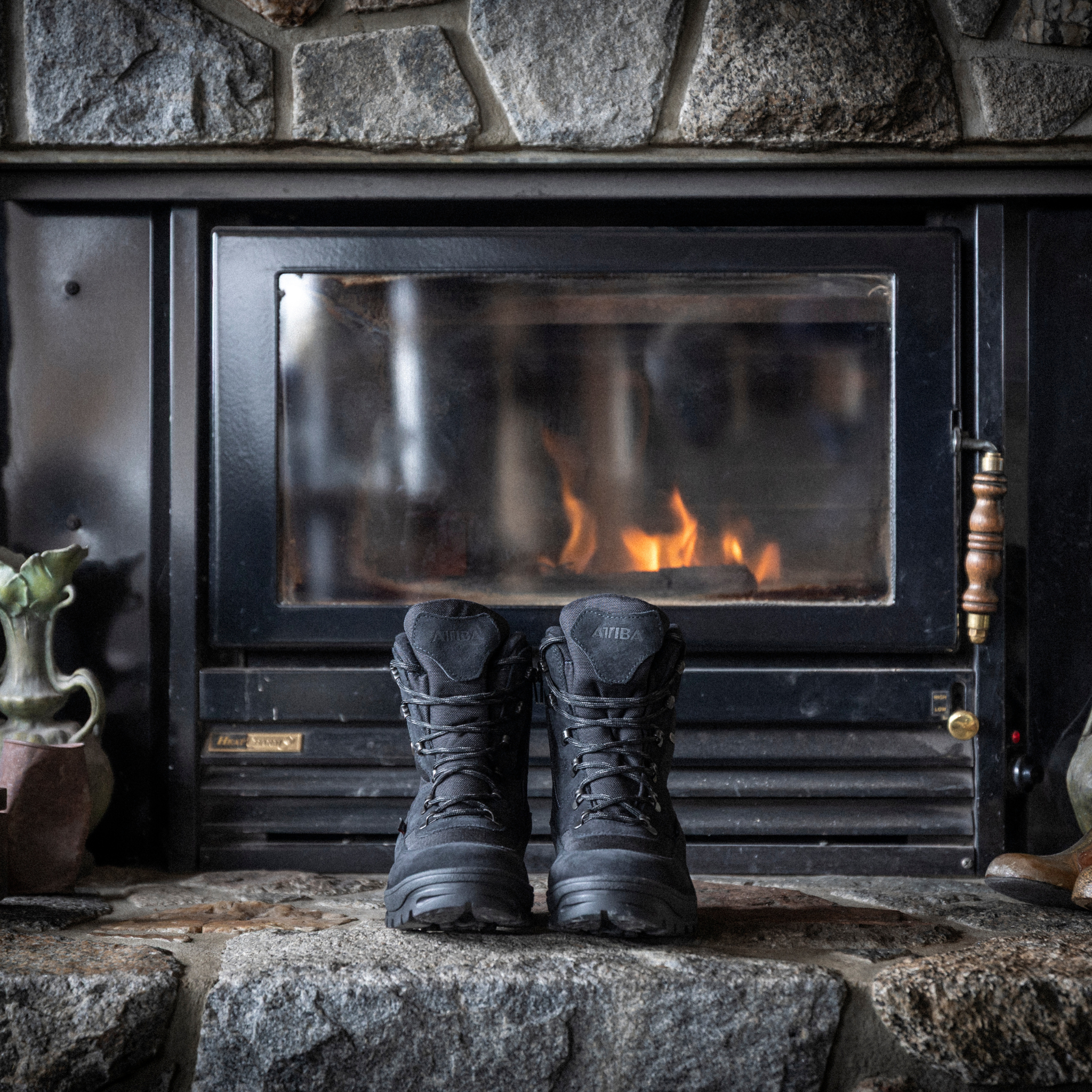 Pair of black Attiba Eiger 2 men's lace-up snow boots warming by a fireplace on a stone hearth, lifestyle view