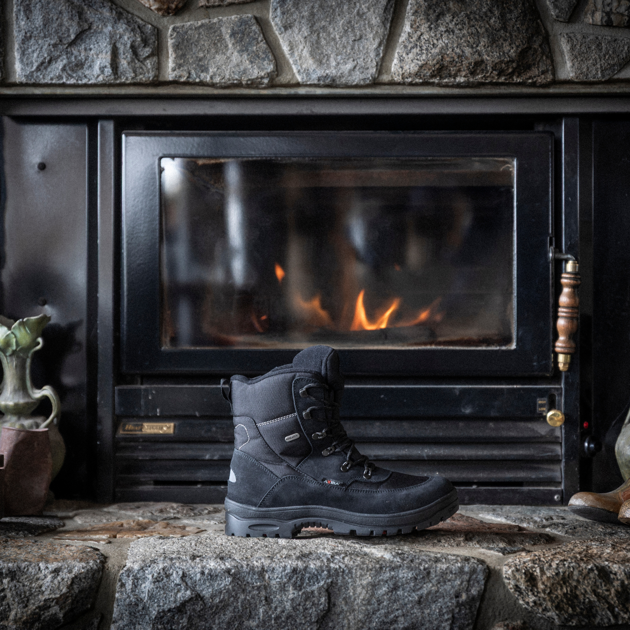 Black Attiba Eiger 2 men's lace-up snow boot on a stone hearth beside a lit fireplace, lifestyle view